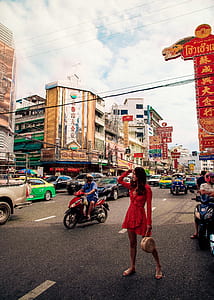 photo of woman wearing red dress holding bag standing on asphalt road surrounded by vehicles thumbnail
