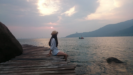 woman wearing white long-sleeved top sitting on brown wooden dock facing body of water thumbnail