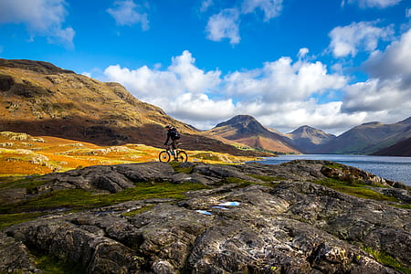 person riding on mountain bike with backpack on rocky mountain near body of water under blue sky at daytime thumbnail