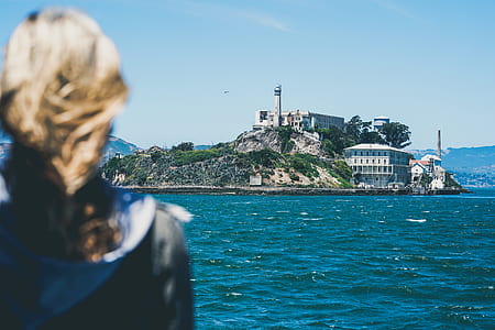 blonde haired woman standing in front of large body of water near island with lighthouse tower during daytime thumbnail