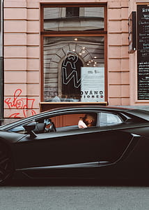 matte-black Lamborghini Aventador with man inside parked beside beige concrete building thumbnail