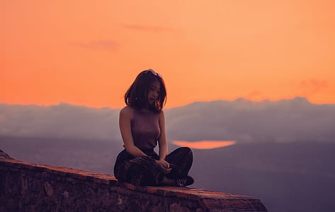 woman in purple sleeveless top and black pants sitting on the edge of cliff at sunset thumbnail