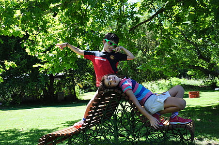 boy standing on bench beside girl lying on backrest under shade of tree on park at daytime thumbnail