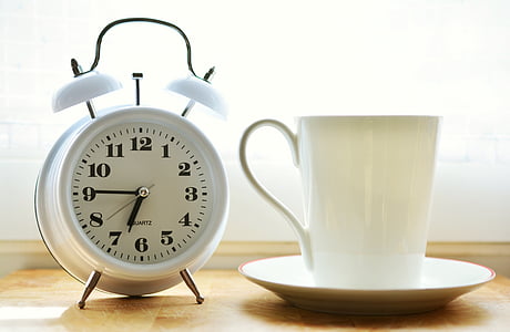 closeup photo of white ceramic mug on saucer beside white twin-bell alarm desk clock on brown wooden table thumbnail