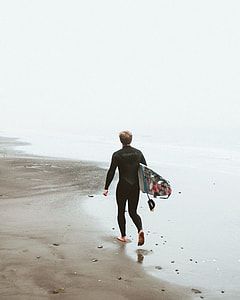 man wearing wet suit walking on seashore carrying surfing board thumbnail