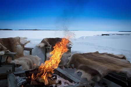 burning bonfire beside chairs with animal skin pads surrounded by snow during daytime thumbnail