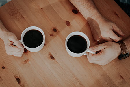 two person's holding white ceramic mugs on brown table thumbnail