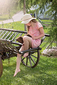 girl wearing white hat and pink floral short-sleeved dress sitting on brown wooden trailer while holding book thumbnail