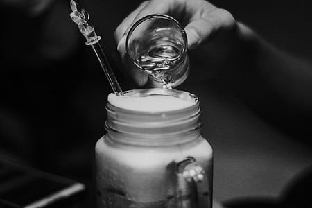 grayscale photo of person holding shot glass pouring beverage on beverage in glass mason jar thumbnail