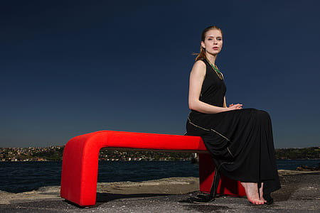 woman in black sleeveless dress sitting on red bench near body of water thumbnail