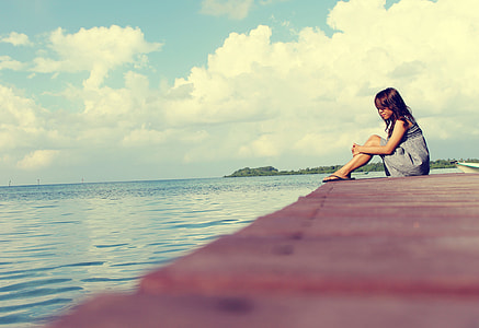 woman in gray dress sitting at dock area facing body of water thumbnail