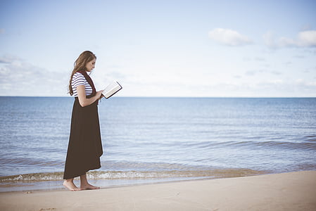 woman holding book while standing at seashore during daytime thumbnail