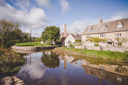 beige concrete house surrounded with green grass field near river under white clouds and blue sky thumbnail