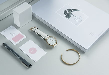 round gold-colored and white analog watch with white leather band beside black pen and white labeled box thumbnail