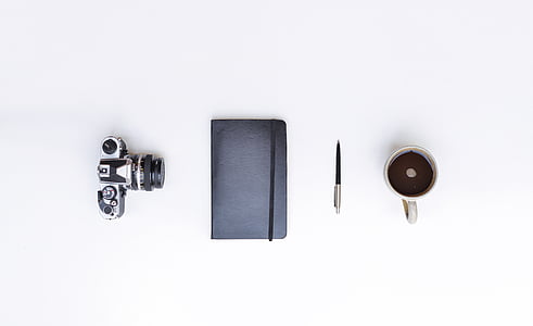 photo of DSLR camera, black handbook, pen, and mug filled with coffee on white surface thumbnail