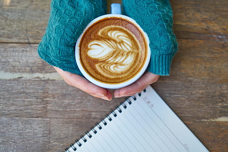 person holding heart cappuccino with both hands near white springside lined notebook above wooden surface thumbnail