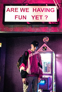 man wearing multicolored jacket standing near doll claw machine with siganage thumbnail