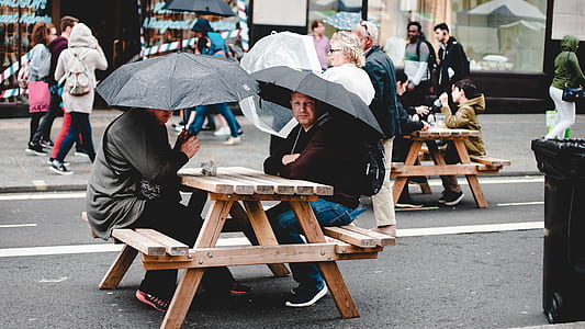 two men sitting on brown wooden picnic table while using umbrellas thumbnail