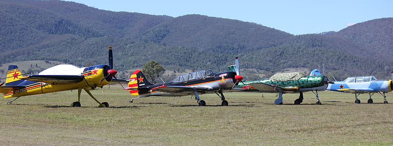 four assorted-colored airplanes on green grass field near green mountain under blue sky thumbnail