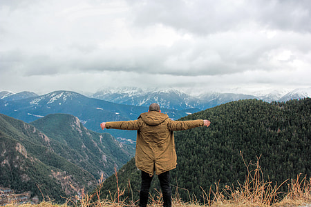 man wearing jacket standing on brown grass at the hill facing glacier mountains under white clouds during daytime thumbnail
