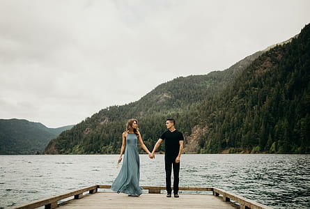 man wearing black v-neck shirt beside woman wearing green maxi dress standing on brown wooden boat dock during day time thumbnail