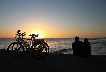 silhouette photography of two person sitting beach shore beside bicycles thumbnail