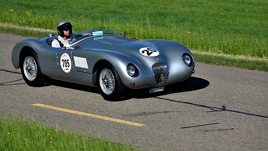 grey classic racing roadster on concrete track driven by helmet wearing driver thumbnail