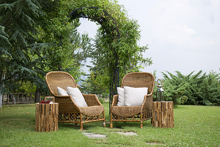 two brown wicker armchairs with white throw pillows beside two round tables on grass field thumbnail