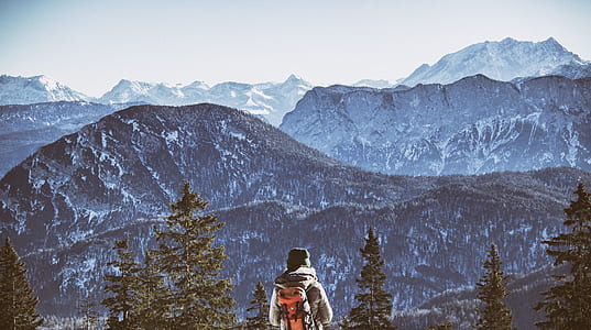 person with backpack standing on top of mountain with trees during day time thumbnail
