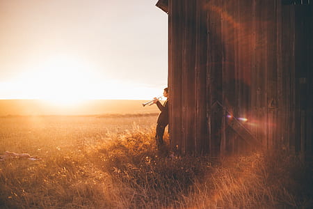 person holding trombone while leaning on brown wooden house during golden hour thumbnail