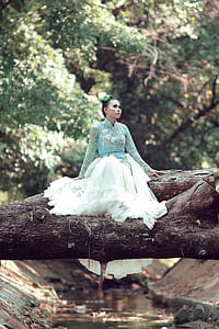 woman in blue and white floral long-sleeved dress sitting on brown tree trunk over body of water during daytime thumbnail