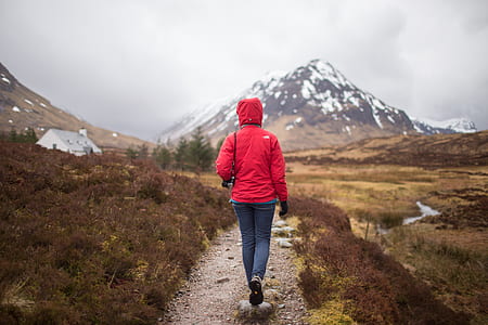 person in red hooded jacket and blue denim jeans walking near mountain covered by snow during daytime thumbnail