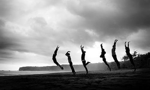 grayscale photography of six people jumping at sand nearby body of water thumbnail