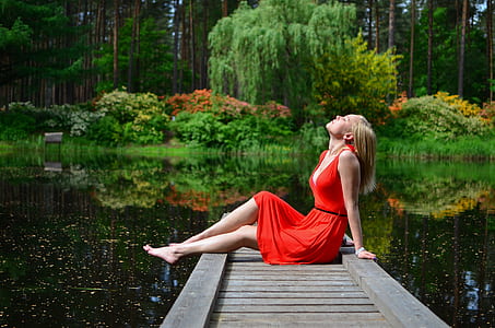 photo of woman sitting near edge of dock surrounded by body of water far away from tall trees thumbnail