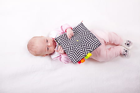 baby wearing pink pajama sleeper lying on white comforter and holding black cushion thumbnail