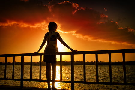 silhouette of woman holding on hand rail in front of body of water at golden hour thumbnail