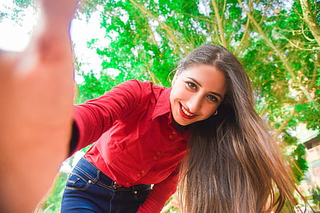 Smiling Woman in Red Shirt and Blue Jeans Taking Selfie Under Green Leaved Tree thumbnail