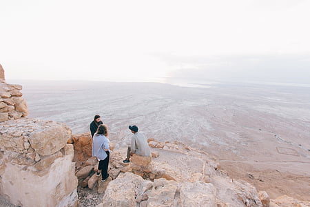 three men standing and sitting on big rock under white sky during daytime thumbnail