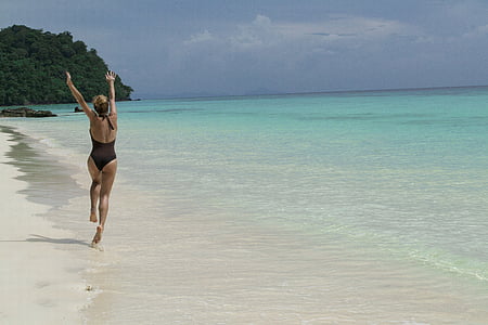 woman wearing one-piece swimsuit doing hands up while running on beach shore thumbnail