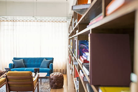 tufted blue suede couch beside brown leather ottoman and brown wooden bookshelf inside room thumbnail
