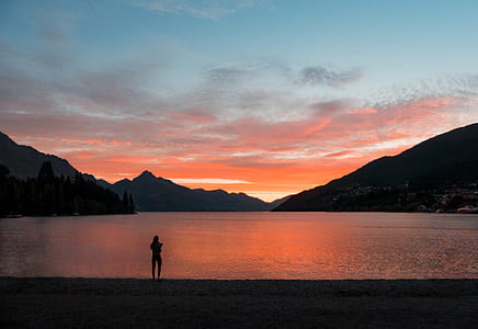 silhouette of person standing at the seashore across mountains photo during golden hour thumbnail