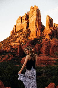 woman wearing black sleeveless top and brown skirt standing near rock formation overlooking brown mountain thumbnail