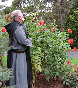 man in grey robe standing near red flowers during daytime thumbnail