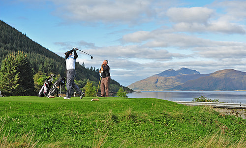 man in white shirt playing golf on green field beside man in black shirt under white cloudy sky thumbnail