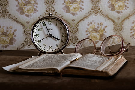 round gray analog desk clock beside eyeglasses and opened book on brown wooden surface thumbnail