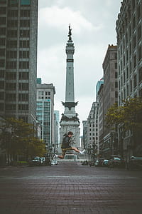 man in black hooded jacket jumping on mid-air in front of white statue between commercial high-rise building during daytime shallow focus photography thumbnail