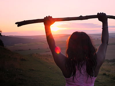 woman raising two hands holding brown wood facing sunset thumbnail