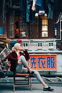 woman in red fur jacket and blue jeans sitting on wooden folding chair in front of brown house with hanging clothes during daytime thumbnail