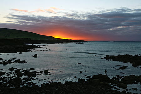 silhouette of man standing on rocks facing body of water thumbnail