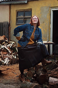 woman wearing blue denim button-up coat and black skirt standing with right hand on hip and left foot on brown firewood thumbnail